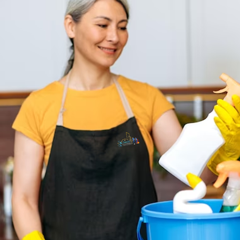 A woman in an apron and gloves is holding a bucket of cleaning supplies.